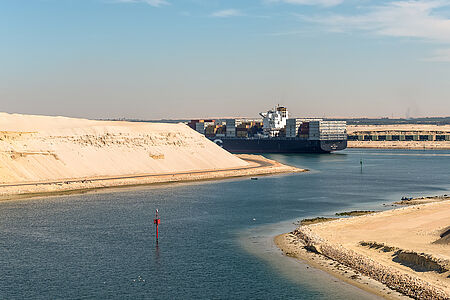 Ein großes Containerschiff fährt durch einen breiten Kanal in einer sandigen Wüstenlandschaft unter blauem Himmel.